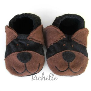 May include: A pair of baby booties in a dog design. The booties are brown and black, with a dog face design on the front. The booties have a soft, felt-like texture and are designed to look like a dog's face. The booties are on a white background.