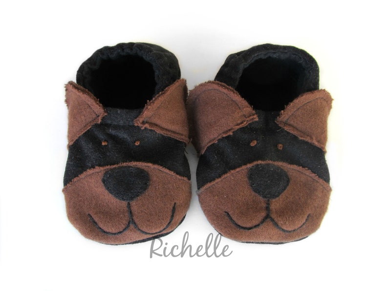 May include: A pair of baby booties in a dog design. The booties are brown and black, with a dog face design on the front. The booties have a soft, felt-like texture and are designed to look like a dog's face. The booties are on a white background.