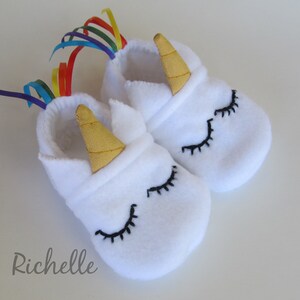 May include: White fleece baby booties with a unicorn design. The booties have a gold horn, black eyelashes, and a rainbow ribbon.