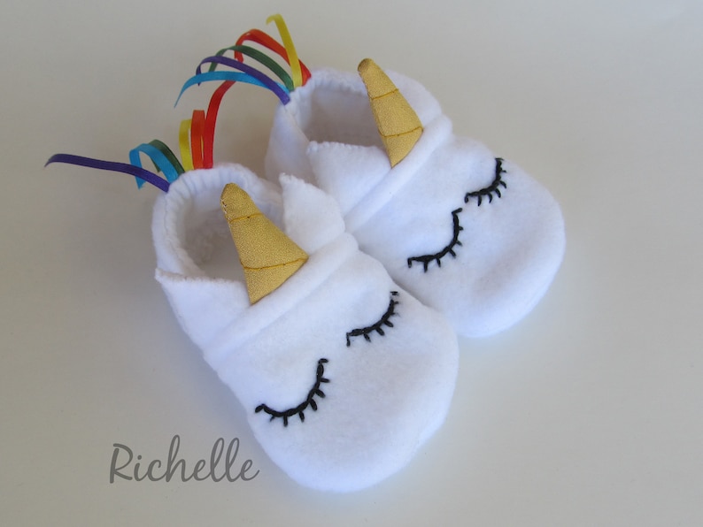 May include: White fleece baby booties with a unicorn design. The booties have a gold horn, black eyelashes, and a rainbow ribbon.