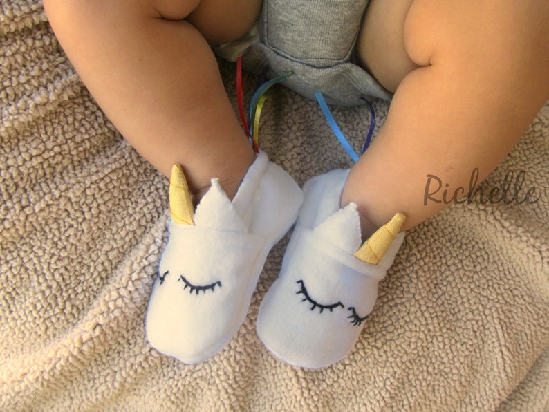 May include: White baby booties with unicorn ears and closed eyes. The booties have a yellow horn and are made of soft, plush fabric.