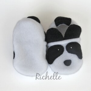 May include: A pair of gray fleece baby booties with black panda bear faces. The booties have black felt ears and eyes.