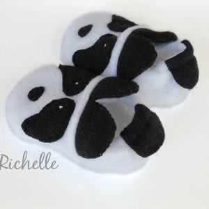 May include: A pair of white baby booties with black felt panda bear faces. The booties are made of soft, plush fabric.