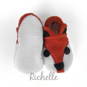 May include: A pair of baby booties with a fox design. The booties are made of soft, brown fleece and have a white felt bottom with a textured pattern. The fox face features black felt eyes, nose, and ears.