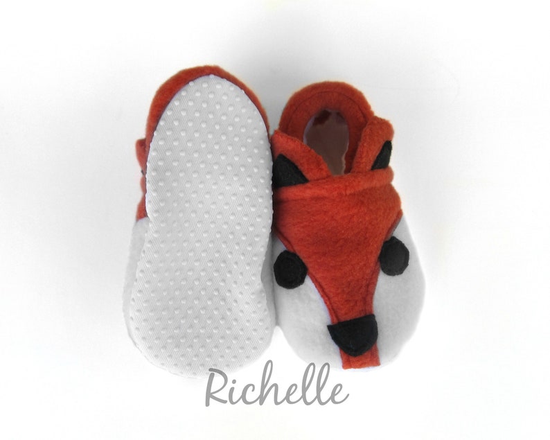 May include: A pair of baby booties with a fox design. The booties are made of soft, brown fleece and have a white felt bottom with a textured pattern. The fox face features black felt eyes, nose, and ears.