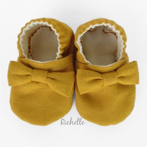 May include: A pair of mustard yellow baby shoes with a bow on the front. The shoes are made of a soft fabric and have a white lining.