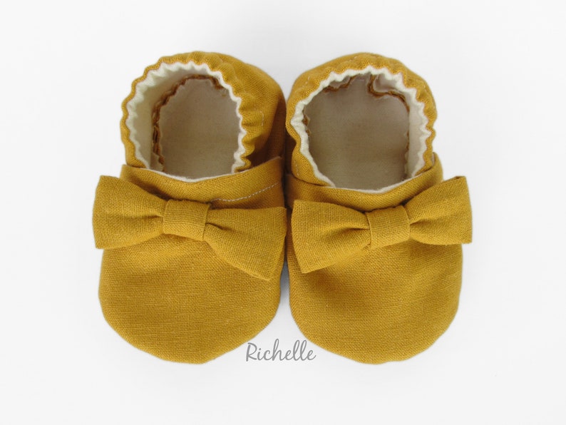 May include: A pair of mustard yellow baby shoes with a bow on the front. The shoes are made of a soft fabric and have a white lining.