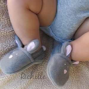 May include: Gray baby booties shaped like bunnies, with pink inner ears and small white accents. The booties are on a baby's feet, with a gray onesie visible. The soft, felt-like material suggests comfort and warmth for infants.