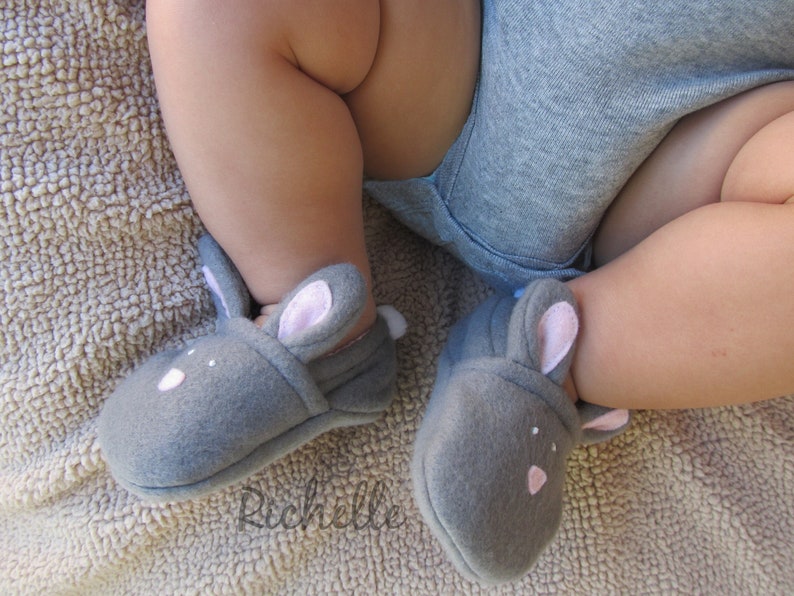 May include: Gray baby booties shaped like bunnies, with pink inner ears and small white accents. The booties are on a baby's feet, with a gray onesie visible. The soft, felt-like material suggests comfort and warmth for infants.