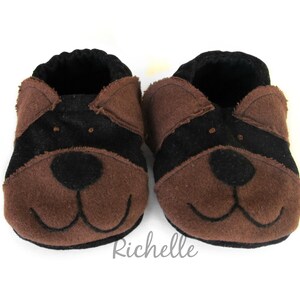 May include: A pair of baby booties in a dog design. The booties are brown and black, with a black stripe across the top and a dog face on the front. The booties are made of soft material and have a black interior.