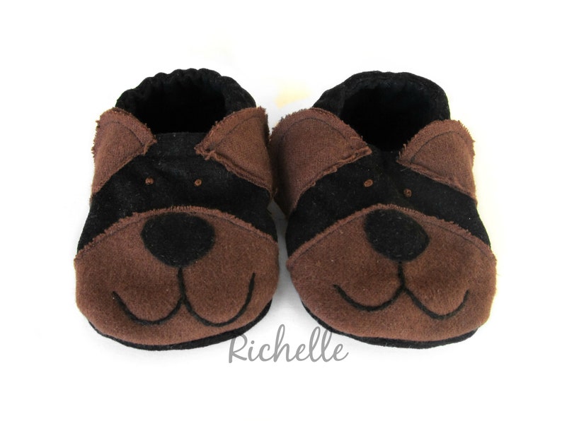 May include: A pair of baby booties in a dog design. The booties are brown and black, with a black stripe across the top and a dog face on the front. The booties are made of soft material and have a black interior.