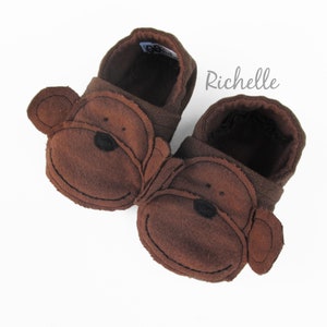 May include: A pair of brown fleece baby booties with monkey faces. The booties have brown felt ears and a black nose.