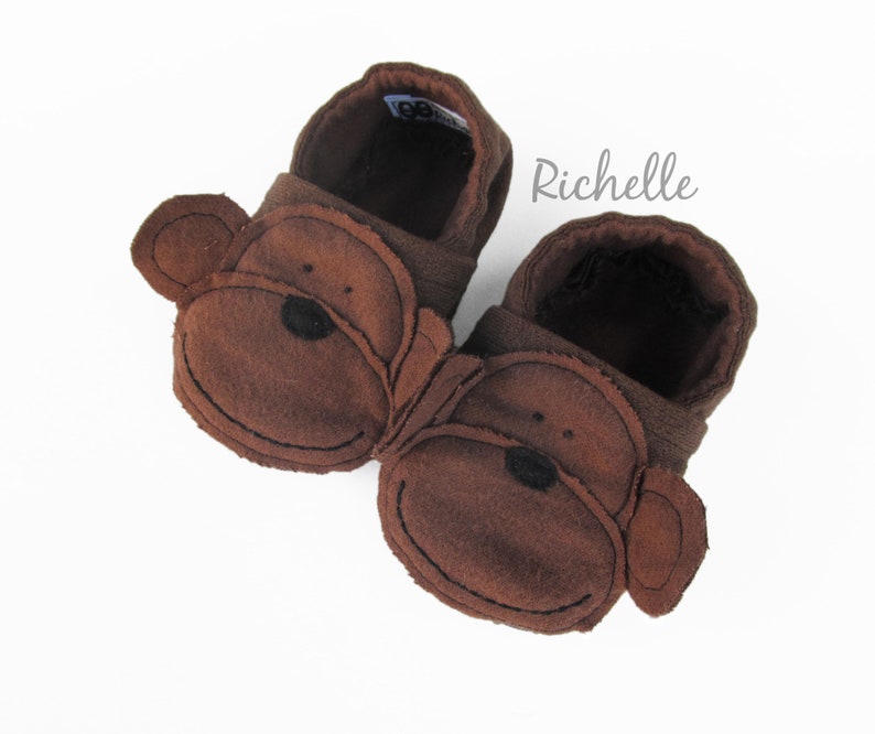 May include: A pair of brown fleece baby booties with monkey faces. The booties have brown felt ears and a black nose.