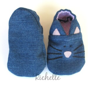 May include: A pair of blue denim baby shoes with a cat face design. The cat face features black whiskers, a pink nose, and pink ears.