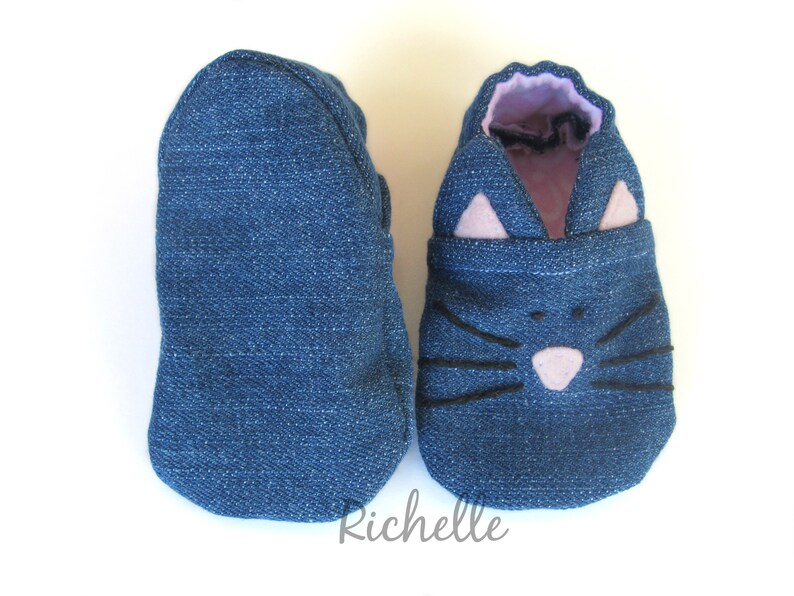 May include: A pair of blue denim baby shoes with a cat face design. The cat face features black whiskers, a pink nose, and pink ears.