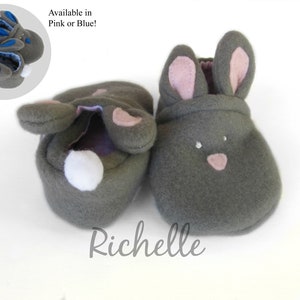 May include: Gray bunny-shaped baby booties with pink inner ears and nose details. A white pom-pom tail is on the back of one bootie. The image includes a smaller inset showing blue accents. The text "Available in Pink or Blue!" is present.
