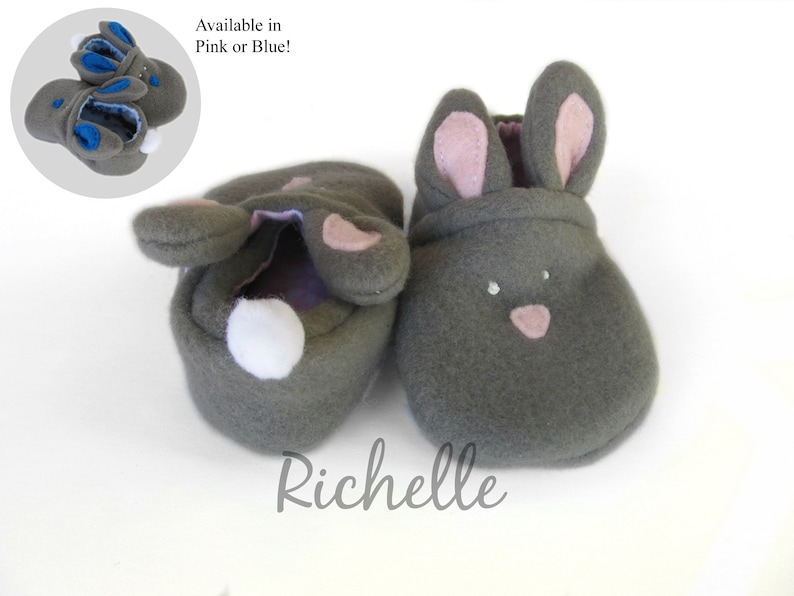 May include: Gray bunny-shaped baby booties with pink inner ears and nose details. A white pom-pom tail is on the back of one bootie. The image includes a smaller inset showing blue accents. The text "Available in Pink or Blue!" is present.