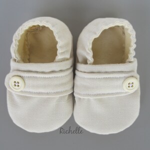 May include: A pair of beige baby booties with a button closure. The booties are made of a soft, textured fabric.