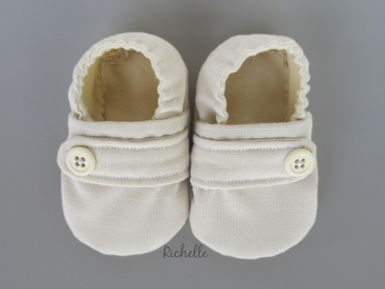 May include: A pair of beige baby booties with a button closure. The booties are made of a soft, textured fabric.