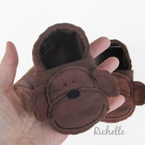 May include: A pair of brown baby booties with a monkey face design. The booties are made of brown fabric with a stitched-on monkey face featuring black eyes and a black nose.
