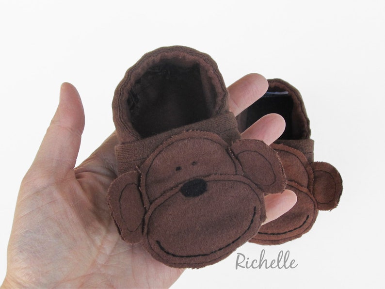 May include: A pair of brown baby booties with a monkey face design. The booties are made of brown fabric with a stitched-on monkey face featuring black eyes and a black nose.