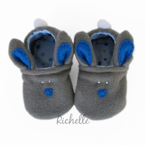 May include: A pair of gray baby booties shaped like bunnies. The booties have blue inner ears and noses, white pom-poms, and a blue polka-dot lining. The booties are designed to look like a bunny.