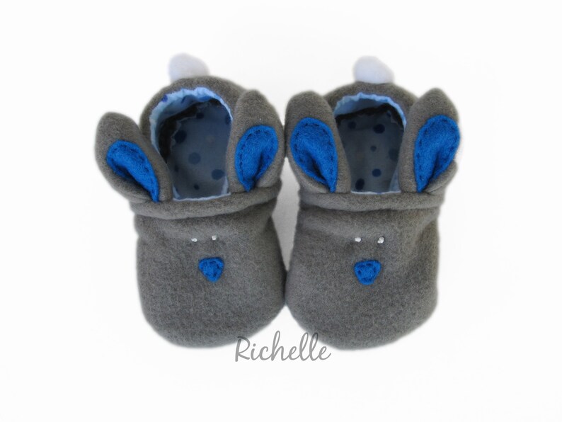 May include: A pair of gray baby booties shaped like bunnies. The booties have blue inner ears and noses, white pom-poms, and a blue polka-dot lining. The booties are designed to look like a bunny.