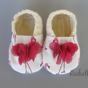 May include: A pair of white baby shoes with a pink flamingo print and red fabric flower accents.