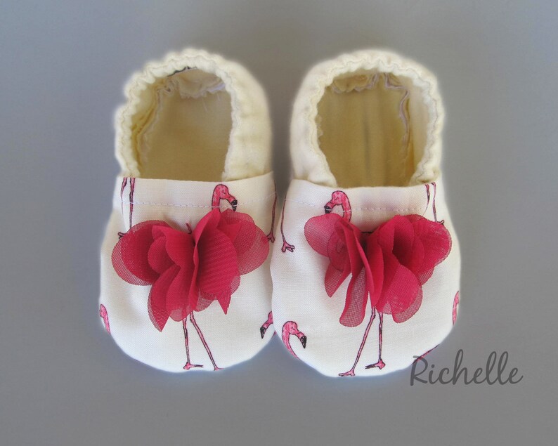 May include: A pair of white baby shoes with a pink flamingo print and red fabric flower accents.