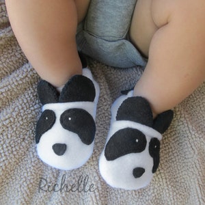 May include: A pair of white and black panda baby booties with black felt ears and eyes. The booties are on a brown fuzzy blanket.
