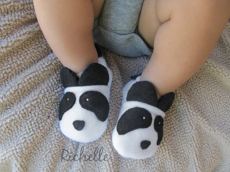 May include: A pair of white and black panda baby booties with black felt ears and eyes. The booties are on a brown fuzzy blanket.
