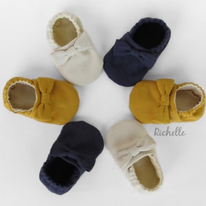 May include: Six pairs of baby shoes in various colours, including beige, mustard yellow, and navy blue. Each shoe has a bow on the top. The shoes are arranged in a star shape on a white background. The word "Richelle" is printed on the background.