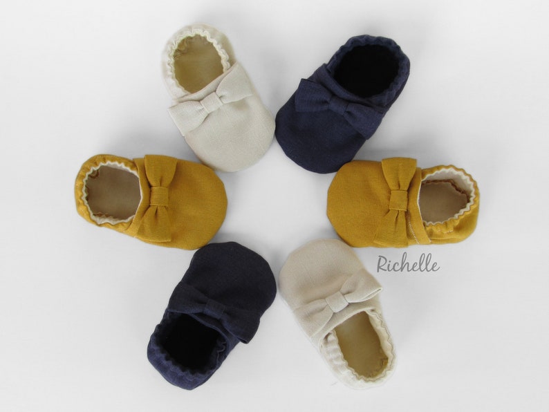 May include: Six pairs of baby shoes in various colours, including beige, mustard yellow, and navy blue. Each shoe has a bow on the top. The shoes are arranged in a star shape on a white background. The word "Richelle" is printed on the background.