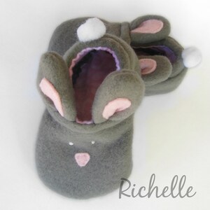 May include: Gray felt bunny slippers with pink inner ears and nose, and white pom-poms. The slippers have a soft, plush appearance and are designed to resemble a bunny rabbit. The slippers are handmade and feature a cute, whimsical design.