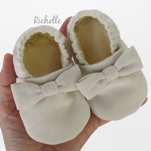 May include: A pair of beige baby shoes with a bow on the front. The shoes are made of a soft, textured fabric.