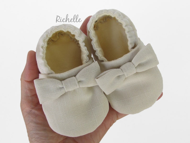 May include: A pair of beige baby shoes with a bow on the front. The shoes are made of a soft, textured fabric.