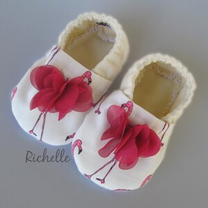 May include: A pair of white baby shoes with a flamingo print and red fabric flowers. The shoes have a soft, cream-coloured lining.