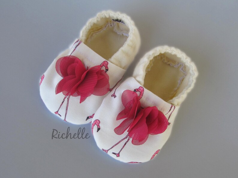 May include: A pair of white baby shoes with a flamingo print and red fabric flowers. The shoes have a soft, cream-coloured lining.