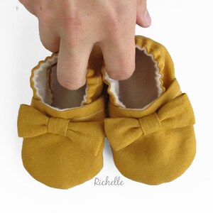 May include: A pair of mustard yellow baby shoes with a bow on the front. The shoes are made of a soft, linen fabric.