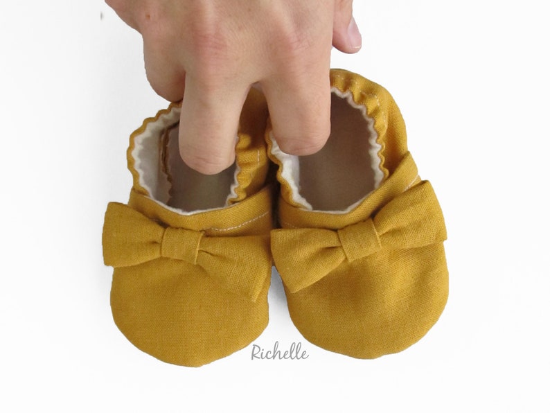 May include: A pair of mustard yellow baby shoes with a bow on the front. The shoes are made of a soft, linen fabric.