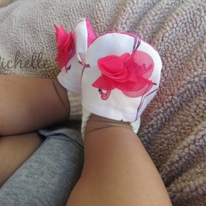 May include: A pair of white baby booties with pink flamingo designs and pink flower accents. The booties are on a baby's legs.