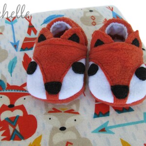 May include: A pair of orange fox baby booties with black eyes and nose. The booties are on a white background with a pattern of foxes, arrows, and teepees.