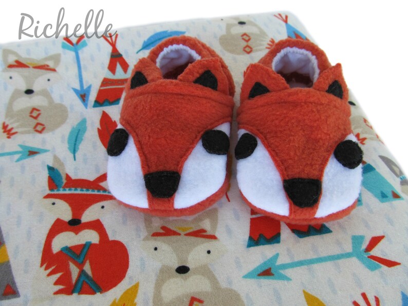 May include: A pair of orange fox baby booties with black eyes and nose. The booties are on a white background with a pattern of foxes, arrows, and teepees.
