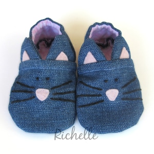 May include: A pair of blue denim baby shoes with a cat face design. The shoes have pink felt ears and a black felt nose and whiskers.