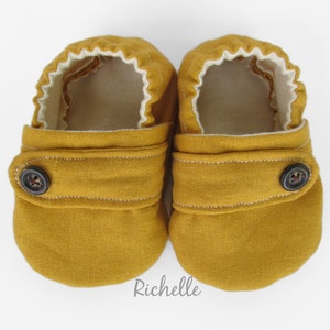 May include: A pair of mustard yellow baby shoes with a button closure and white lining. The shoes are made of a soft, comfortable fabric.