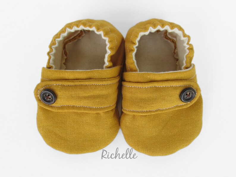 May include: A pair of mustard yellow baby shoes with a button closure and white lining. The shoes are made of a soft, comfortable fabric.
