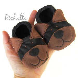 May include: A pair of baby booties in the shape of a dog's face. The booties are brown and black, with embroidered facial features. The word "Richelle" is written in cursive on the upper left side.