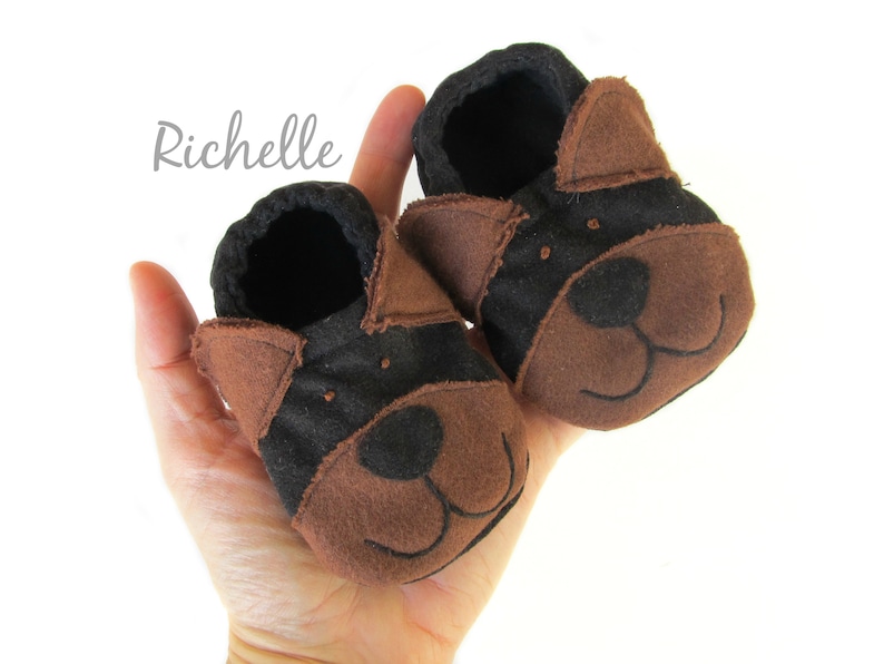 May include: A pair of baby booties in the shape of a dog's face. The booties are brown and black, with embroidered facial features. The word "Richelle" is written in cursive on the upper left side.