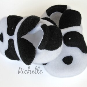 May include: A pair of white fleece baby booties with black felt panda bear faces. The booties have black felt ears, eyes, and noses.