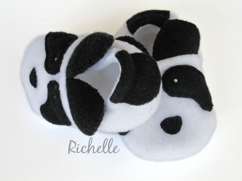 May include: A pair of white fleece baby booties with black felt panda bear faces. The booties have black felt ears, eyes, and noses.